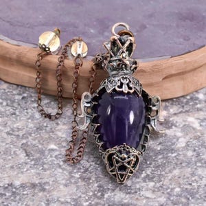 Puede incluir: Colgante plateado con una gran gema púrpura en forma de lágrima. El colgante tiene detalles de filigrana ornamentados y una estrella de cinco puntas en la parte inferior. Una cadena con dos cuentas decorativas y una cadena de color cobre está adjunta.