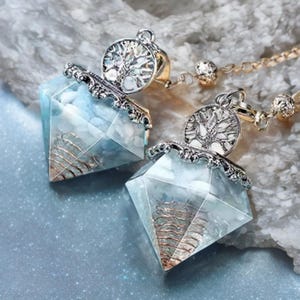 May include: Two diamond-shaped pendants with a light blue hue and a silver-coloured tree of life design. The pendants have a wire-like structure inside and are suspended from a gold-coloured chain.