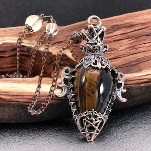 Könnte beinhalten: Eine Halskette mit Anhänger, die einen Tigeraugenstein in einer verzierten, silberfarbenen Metalleinfassung zeigt. Der Anhänger hat eine kronenartige Spitze und ein sternförmiges Design am Boden. Die Kette ist dunkelbronze, der Hintergrund ist Holz. Der Anhänger ist ca. 5 cm lang.