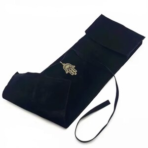 May include: Black velvet jewelry roll with a silver-toned Hamsa hand pendant. The roll is open, revealing a pocket for jewelry storage. A black ribbon is attached for securing the roll.