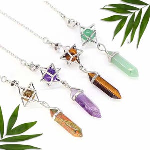 Puede incluir: Una colección de collares plateados, cada uno con un colgante de cristal de diferente color. Los colgantes incluyen cristales verdes, morados, naranjas y marrones, suspendidos de una jaula en forma de estrella. Los collares se exhiben sobre un fondo blanco con hojas verdes.