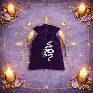 May include: A deep purple velvet drawstring pouch featuring a white graphic print of a snake with celestial moon and star patterns on its body. The bag has a gathered top closure with matching purple fabric cords for securing the contents inside.