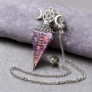 May include: A conical purple crystal pendulum wrapped in a copper wire coil. The top features a silver-toned metal cap with a pentagram symbol flanked by two crescent moons. A silver-toned metal chain with a small decorative bead is attached.