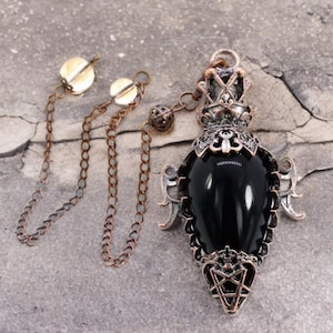 Könnte beinhalten: Ein schwarzer Edelstein-Anhänger mit einem detaillierten silbernen und kupfernen Rahmen. Der Anhänger ist tropfenförmig und hat unten ein Pentagramm-Design. Er ist an einer Kupferkette mit zwei Kupferverschlüssen befestigt.