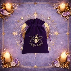 May include: A deep purple velvet drawstring pouch featuring a gold-coloured graphic of a bee with a crescent moon above its head and floral sprigs below. The pouch has matching purple drawstrings and is centred on a purple fabric surface.