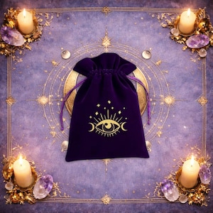 May include: A deep purple velvet drawstring pouch featuring a gold-coloured graphic print of an eye surrounded by crescent moons and stars. The pouch is centred, with a smooth fabric texture and matching purple cord ties at the top closure.