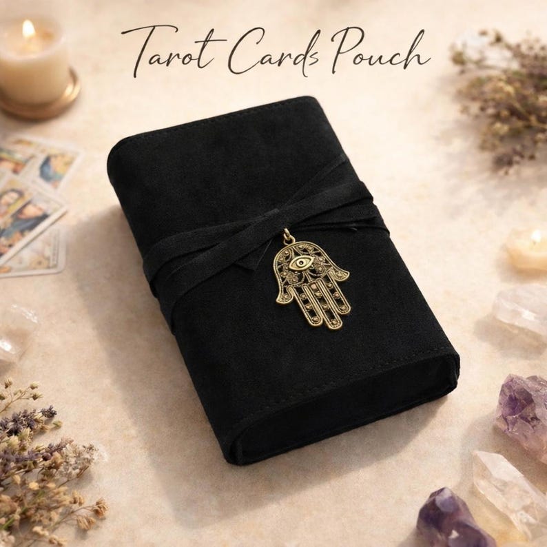 May include: Black tarot card pouch with a wrap-around tie closure and a gold-colored Hamsa hand charm. The pouch has the words "Tarot Cards Pouch" printed on the top. The pouch is made of a soft, suede-like material.