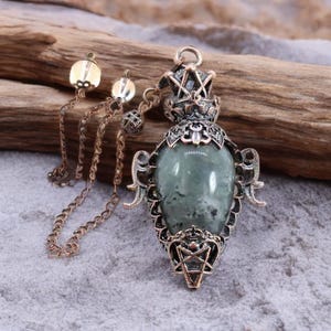 Op de afbeelding: Een vintage-stijl hangerketting met een grote, ovale, groene edelsteen. De hanger is ingekapseld in sierlijk, bronskleurig metaalwerk met een ster aan de onderkant. De ketting bevat een ketting en twee bungelende decoratieve elementen.