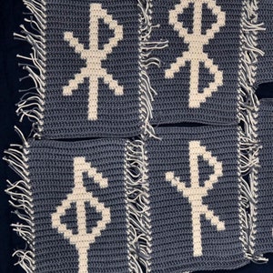 Può includere: Otto quadrati all'uncinetto con frange bianche e rune ricamate bianche su uno sfondo blu.