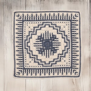 Puede incluir: Un cojín cuadrado de crochet con un patrón geométrico en tonos crema y gris. El cojín tiene un borde gris oscuro y un diseño central en forma de estrella.