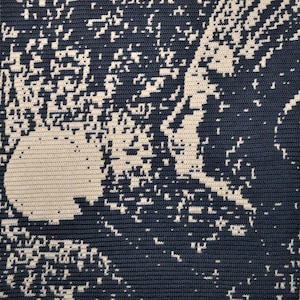May include: A navy blue and cream crocheted blanket with a square shape. The design is an abstract depiction of a celestial scene, with a large cream-coloured sphere and scattered light-coloured shapes against a dark background.