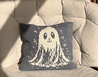 May include: A square crochet pillow in slate gray featuring a centered off-white ghost graphic. The ghost has two dark eyes and a small mouth, surrounded by small white pixelated sparkles. The pillow has a visible textured yarn stitch pattern.
