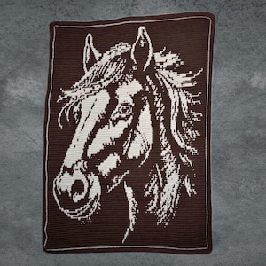 Puede incluir: Una manta de ganchillo marrón y blanca con un diseño de cabeza de caballo. La cabeza del caballo está representada en blanco sobre un fondo marrón, con un borde blanco. La manta muestra un retrato equino detallado.