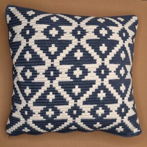 May include: Square crochet pillow with a geometric pattern in navy blue and cream. The pillow features a repeating diamond and cross design, creating a textured surface. The pillow is a decorative home accent.