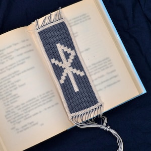 Può includere: Un segnalibro all'uncinetto con un simbolo bianco su sfondo blu scuro. Il segnalibro ha una frangia sul fondo e un lungo fiocco.