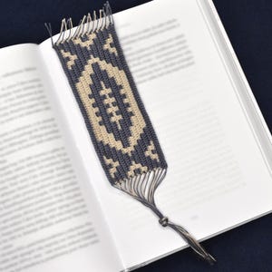 Puede incluir: Un marcapáginas de ganchillo con un patrón geométrico en beige y azul oscuro. El marcapáginas tiene flecos en la parte superior y una borla en la parte inferior.