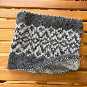 Könnte beinhalten: Ein gestrickter Schlauchschal mit einer dunkel anthrazitfarbenen Basis und einem sich wiederholenden hellgrauen geometrischen Rautenmuster. Das Accessoire hat eine dicke, texturierte Strickstruktur mit einem sichtbaren Innenfutter in einem helleren Grauton.