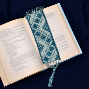 May include: A green and white crocheted bookmark with a tassel. The bookmark is laying on a book.