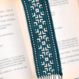 Puede incluir: Un marcapáginas de ganchillo azul turquesa y blanco con un patrón geométrico. El marcapáginas tiene una franja en la parte inferior.
