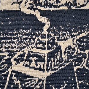 May include: A close-up of a crocheted textile with an abstract design. The piece is mainly navy blue with an off-white pattern. The design resembles a stylized landscape or architectural rendering. The texture is clearly visible.
