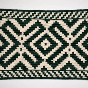 May include: A handmade crochet blanket featuring a geometric pattern in dark green and cream. The design includes diamond and zig-zag shapes, creating a textured and visually appealing textile. The blanket is likely made of soft yarn.