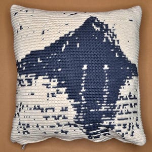 May include: A cream-coloured crochet cushion featuring a large, abstract navy blue design. The design has a pixelated effect, creating a modern, geometric appearance. The cushion has a textured surface and a simple, clean aesthetic.