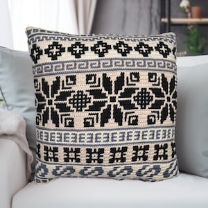 May include: A beige crocheted cushion with a black and grey geometric pattern. The cushion is sitting on a white sofa.