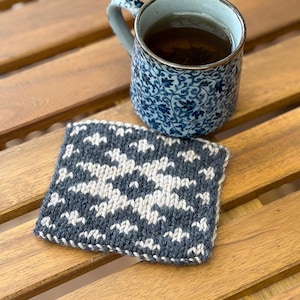Op de afbeelding: Een vierkante gebreide onderzetter met een geometrisch sterpatroon in donkergrijs en crème. Naast de onderzetter staat een keramische mok met een blauw-wit rankenmotief, gevuld met een donkerbruine vloeibare drank.