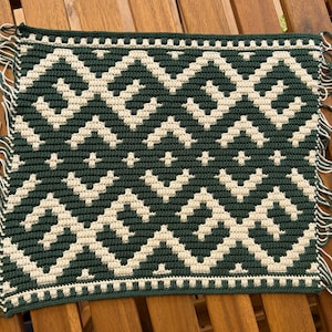Puede incluir: Un textil de ganchillo cuadrado con un patrón geométrico en verde bosque oscuro y crema. El diseño consiste en formas repetitivas de zigzag y diamante con pequeños motivos de cruz. Los bordes están terminados con flecos de hilo verde y crema.