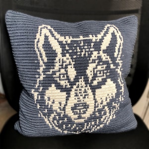 Op de afbeelding: Een vierkant gehaakt kussen in een gedempte blauwe kleur. Het kussen heeft een gedetailleerd, gepixeld ontwerp van een wolfsgezicht in off-white garen. De haaksteken zijn zichtbaar en geven textuur aan het decoratieve kussen.