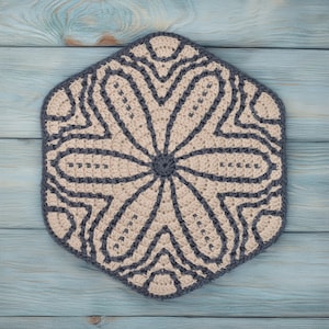 May include: A beige crocheted hexagon-shaped coaster with a gray floral design. The coaster is on a blue wooden surface.