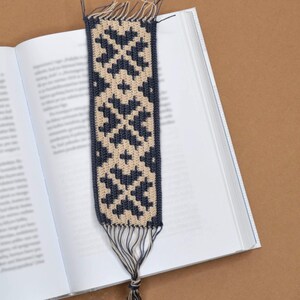 Puede incluir: Un marcapáginas de ganchillo con un patrón geométrico en beige y azul oscuro. El marcapáginas está colgado de un libro.