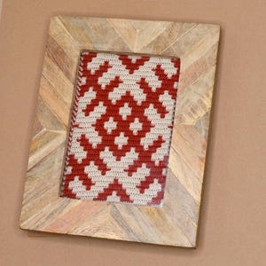 May include: A wooden picture frame with a chevron pattern. The frame holds a red and white crocheted piece of art.