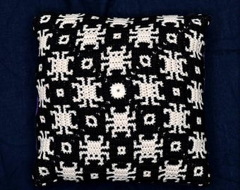 May include: Square black and white crochet pillow with a repeating geometric pattern. The pillow has a dark blue background. The pattern is made of white shapes on a black background. The pillow is square.