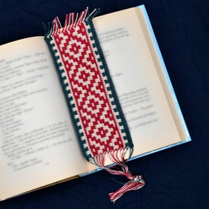 May include: A red, white, and green crocheted bookmark with a fringe tassel. The bookmark is laying on top of a closed book.