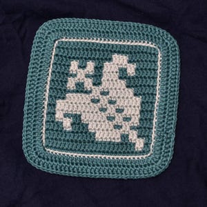 May include: A square, crocheted patch featuring a pixelated design of a video game character. The patch has a teal border with a white inner border, and the character is in shades of gray. The background is a dark blue fabric.