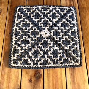 Puede incluir: Funda de cojín cuadrada, tejida a mano, con un patrón geométrico. La funda presenta un fondo azul grisáceo oscuro con un diseño blanco repetitivo. Un pequeño detalle circular está en el centro. La funda mide unos 40 cm cuadrados.