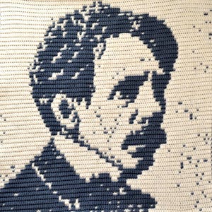 May include: A crocheted portrait in profile, with a pixelated design. The artwork uses navy blue and cream yarn to create a high-contrast image. The piece is likely handmade, showcasing a unique craft technique.