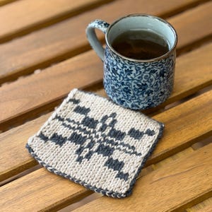 May include: A square, hand-knitted coaster featuring a cream and dark gray geometric pattern with a dark gray border, positioned next to a ceramic mug decorated with a blue and white floral vine motif filled with a dark liquid.