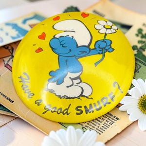 Peut inclure: Un badge rond jaune illustré d'un personnage Schtroumpf bleu portant un bonnet blanc et tenant une marguerite blanche. Le texte "Have a good SMURF?" est imprimé en lettres noires, accompagné de trois petites icônes de cœurs rouges.