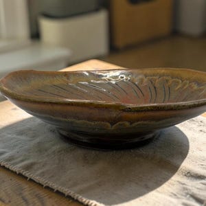 May include: A handcrafted, oval ceramic bowl with a glossy glaze. The bowl has brown, gold, and reddish-orange tones. It sits on a neutral linen cloth, highlighting its unique shape and colour variations. The bowl is likely for serving food.