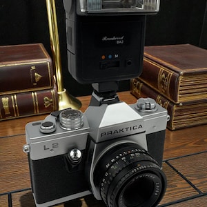 May include: A vintage Praktica L2 camera with a black and silver body, equipped with a lens and a flash unit. The camera is positioned on a wooden surface, with a gold-colored lamp and books in the background.