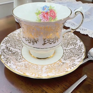 May include: A porcelain teacup and saucer set featuring a pale pink base with intricate gold filigree patterns. The interior of the cup displays a small floral bouquet with pink and blue flowers. The set includes a gold-trimmed handle and rim.