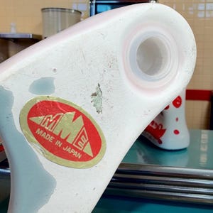 May include: A white ceramic object with a circular opening and a red oval sticker featuring the text MME MADE IN JAPAN. The surface shows signs of wear with some paint loss and a small area of discoloration near the centre of the white base.