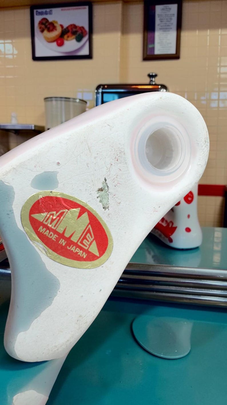 May include: A white ceramic object with a circular opening and a red oval sticker featuring the text MME MADE IN JAPAN. The surface shows signs of wear with some paint loss and a small area of discoloration near the centre of the white base.