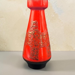 Peut inclure: Un vase en céramique rouge vif avec un col effilé et un bord évasé. Le corps présente un motif en relief texturé avec une finition bronze métallique, tandis que la base est en noir mat uni. Le bord est fini avec un fin liseré doré.