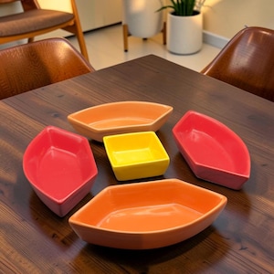 Peut inclure: Un ensemble de cinq plats de service en céramique de formes et de couleurs variées. Il y a un petit bol carré jaune, deux bols allongés orange en forme de bateau et deux bols rouges pentagonaux disposés en groupe sur une surface en bois sombre.