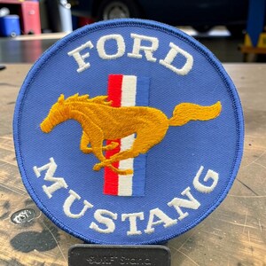 Peut inclure: Un écusson brodé circulaire avec un fond bleu, portant le texte blanc FORD en haut et MUSTANG en bas. Au centre, une silhouette de cheval doré est positionnée sur une barre verticale composée de bandes rouge, blanche et bleue.