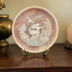 May include: A round decorative plate with a marbled pink and white surface featuring a raised white relief of a figure in a flowing dress, a bird in flight, and surrounding foliage. The plate is supported by a small gold-toned metal stand.