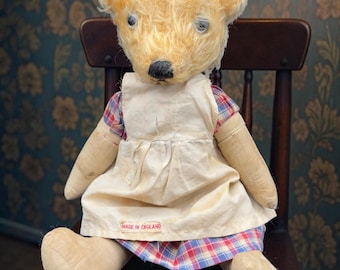 May include: A vintage teddy bear with light brown fur, wearing a cream-colored dress over a plaid skirt. The bear is seated on a wooden chair. The tag on the dress reads "MADE IN ENGLAND".
